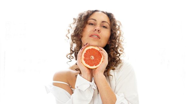 Portrait of pretty middle-aged woman with curly hair with grapefruit at home - white background. The concept of happiness, beauty and health