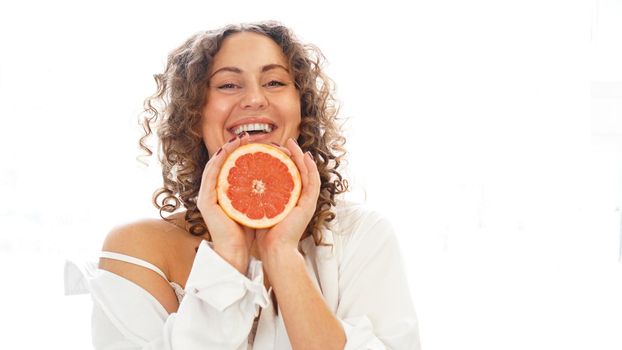 Portrait of pretty middle-aged woman with curly hair with grapefruit at home - white background. The concept of happiness, beauty and health