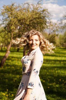 Happy young woman with blonde hair, wearing a dress, posing outdoors in a garden with cherry trees in the sun, smiling. Curls fluttering in the wind