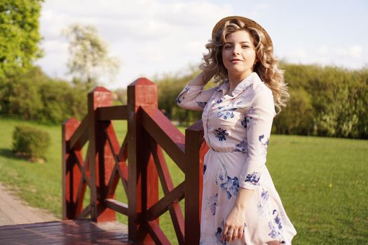 Beautiful woman in a white summer dress and a straw hat stands on a birch bridge in the park