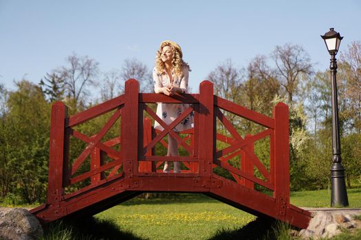 Beautiful woman in a white summer dress and a straw hat stands on a birch bridge in the park