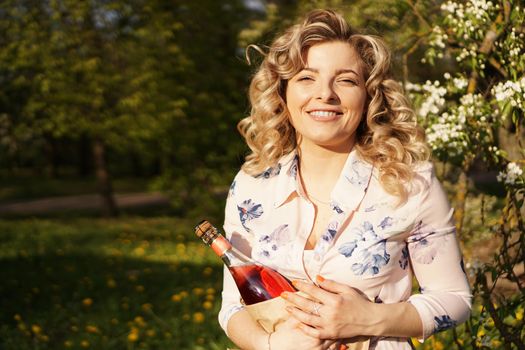 Beautiful young woman holding a blank bottle of wine while having lunch outdoors. Picnic in the park with green grass