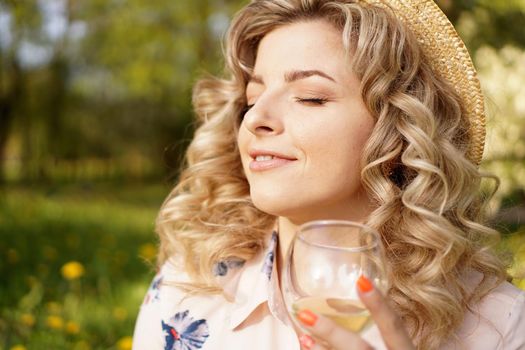 Happy blonde holding a glass of white wine during a picnic at sunset in the green park at summer day