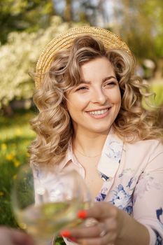 Women clinking glasses with tasty wine on light background at summer day. Happy blonde with curly hair in a straw hat