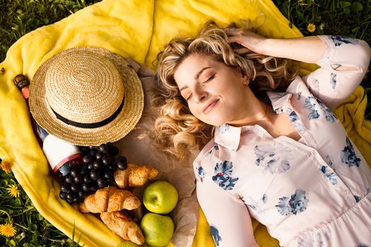 Young girl in hat lying on the plaid with wine and fresh food. Picnic. Natural light