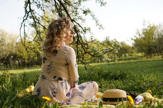 Beautiful blond woman - happy lifestyle, weekend out for a walk in a picnic park in the summer garden, model sits on plaid with food - summer weather