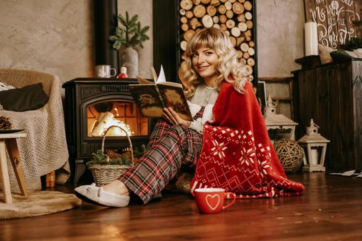 Happy young woman reading book in front of christmas interior with fireplace. The girl is dressed in homely cozy clothes with a red warm plaid