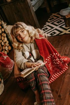 Happy young woman reading book in front of christmas interior with fireplace. The girl is dressed in homely cozy clothes with a red warm plaid