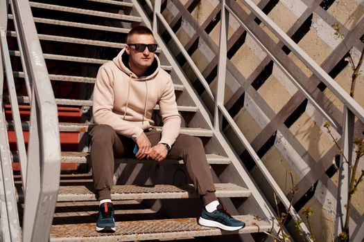 A young man in sunglasses sits on the steps in the city. Urban style. Lifestyle photo