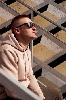 A young man in sunglasses sits on the steps in the city. Urban style. Lifestyle photo. Vertical photo