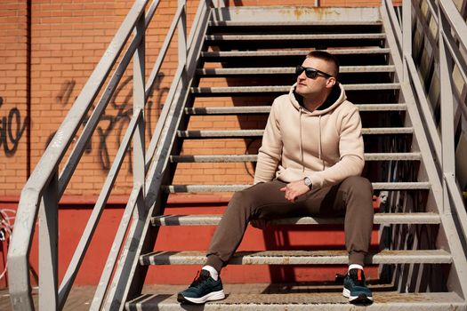 A young man in sunglasses sits on the steps in the city. Urban style. Lifestyle photo