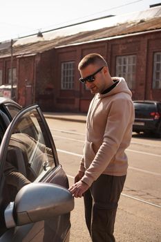 A young man next to his car on a sunny day on the street of the old city. The man opens the door. Lifestyle photo.