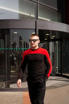A young athletic man in sunglasses in a tracksuit walks out of the revolving doors of a hotel or shopping mall