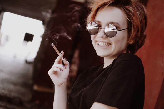 Millennial cool pretty girl with short red hair and mirror sunglasses smoking cigarette in the old city with red walls. Smiling happy teen girl