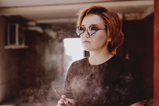 Millennial cool pretty girl with short red hair and mirror sunglasses smoking cigarette in the old city with red walls