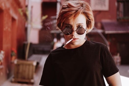 Millennial cool pretty girl with short red hair and mirror sunglasses smoking cigarette in the old city with red walls