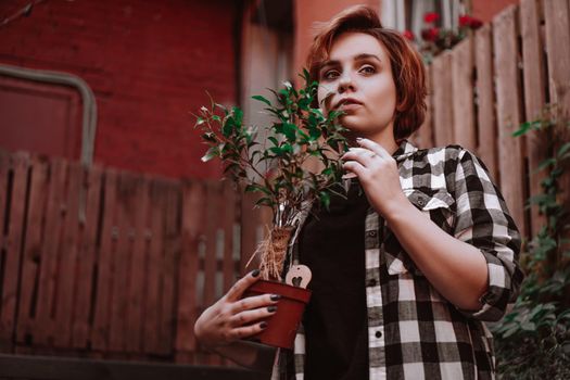 Beautiful young woman with short red hair in a plaid shirt holding a flower in a pot in the backyard