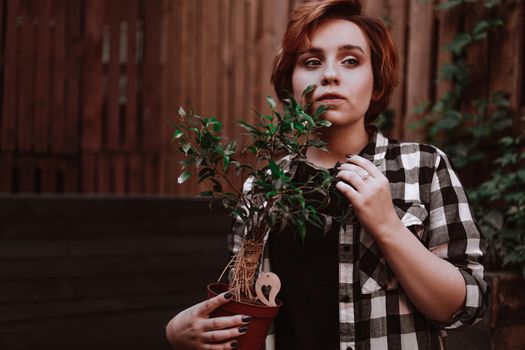 Beautiful young woman with short red hair in a plaid shirt holding a flower in a pot in the backyard