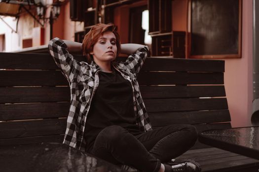 Young woman with short red hair in a plaid shirt in a bar put her legs on the bench