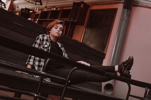 Young woman with short red hair in a plaid shirt in a bar put her legs on table