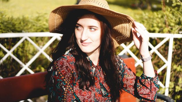 Fashionably dressed young pretty woman on the street on a sunny day. Beautiful girl in dress and hat is sitting on a bench and smiling. Street-fashion concept