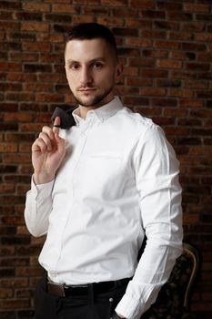 Stylish young businessman in white shirt is holding a jacket on finger, standing against brick wall