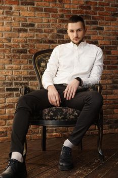 Portrait handsome fashionable man in a white shirt sits on a chair in a photo studio loft. Young businessman is thinking about future