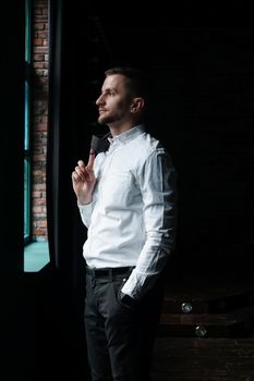 View in profile of a young businessman, dressed in a white shirt standing near the window on a dark walls and looking out the window. Vertical photo