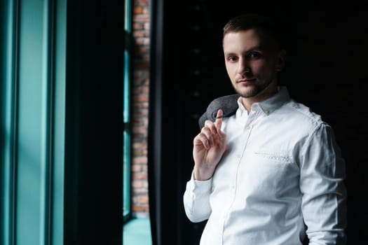 View in profile of a young businessman, dressed in a white shirt standing near the window on a dark walls and looking at the camera