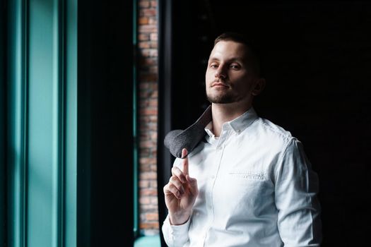 View in profile of a young businessman, dressed in a white shirt standing near the window on a dark walls and looking at the camera