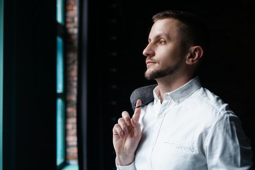 View in profile of a young businessman, dressed in a white shirt standing near the window on a dark walls and looking out the window.