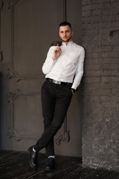 Stylish young businessman in white shirt is holding a jacket on finger, standing against brick wall - Vertical photo