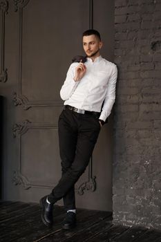 Stylish young businessman in white shirt is holding a jacket on finger, standing against brick wall - Vertical photo