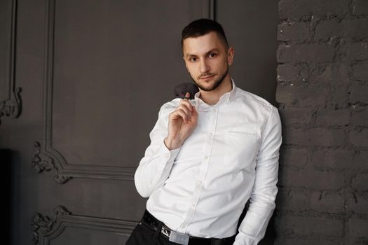 Stylish young businessman in white shirt is holding a jacket on finger, standing against brick wall