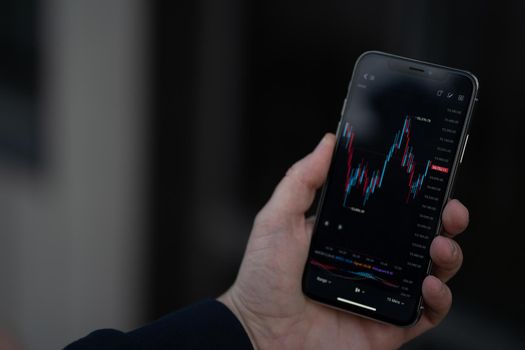 Mobile app for trading online. Male hand holding smartphone with financial forex graph chart on screen, trader or investor checking stock exchange market data while standing outdoors, selective focus