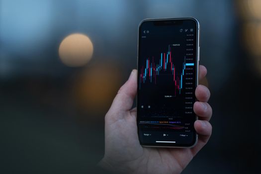 Stock exchange and trading online. Male hand holding smartphone and using investment app, analyzing market data in real time. Selective focus on mobile phone with financial graph chart on screen