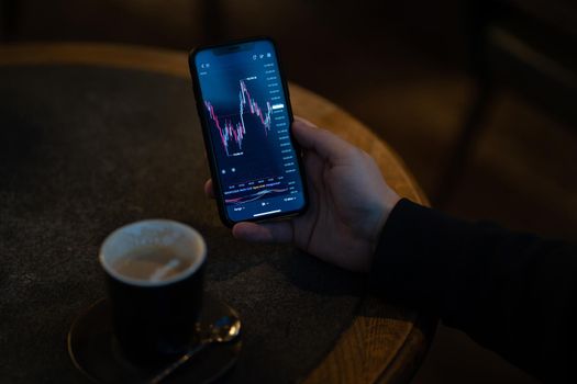 Trader or investor reading financial news and analyzing real time stock market data while sitting outdoors and drinking coffee, male hand holding smartphone with live forex chart on screen