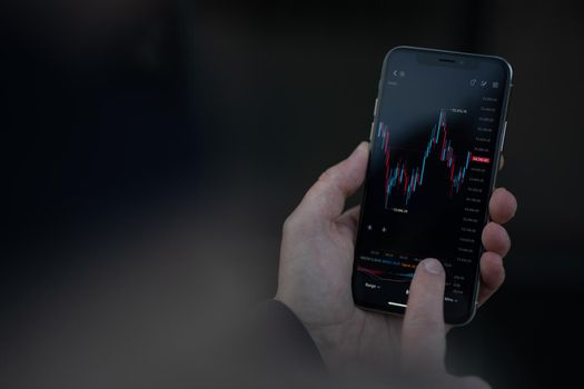 Cropped shot of trader managing investments and trade stocks in mobile app, male hand touching smartphone screen display with financial graph chart. Stock exchange market and trading online concept