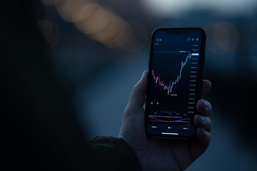 Broker or investor using stock trading app on smartphone to analyzing price flow on stock market while standing outdoors. Selective focus on hand holding mobile phone with forex graph chart on screen