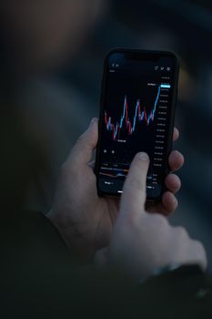 Trader checking stock market data in mobile app for trading online. Male hand touching smartphone screen with real time forex chart, reading financial news and analyzing price flow, selective focus