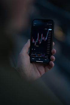 Trading wherever you are. Male hand using mobile app on smartphone for reading financial news in real time and checking stock market data in real time. Selective focus on screen with forex graph chart