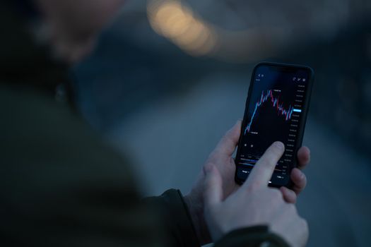 Rear view of trader monitoring stock market data in mobile app for online trading and investing on smartphone while standing outdoors, selective focus on hand touching screen with forex graph chart