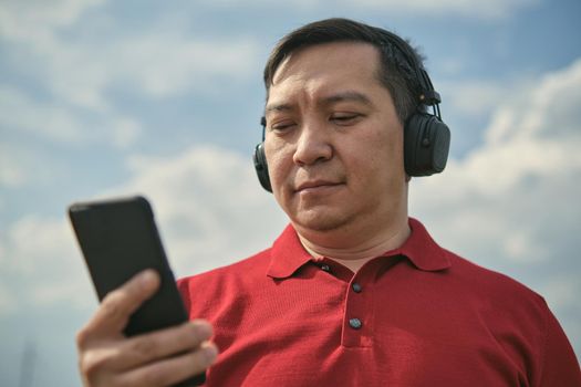 Middle-aged Asian man in headphones outdoors listening to music against the background of the sky, mobile phone in his hand