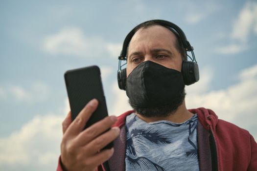 Middle-aged European man in headphones outdoors listening to music against the background of the sky, mobile phone in his hand