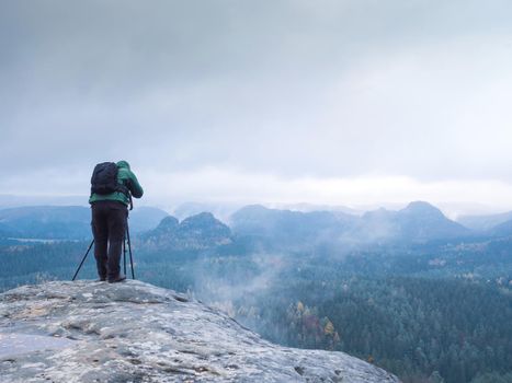 Travel hobby photographer taking picture of misty mountain landscape using professional camera on tripod on stormy, autumn day