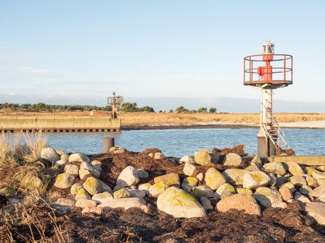 Red and white small technical lighthouse on the stony pier viewed from low angle with dry grass and white stones in foreground