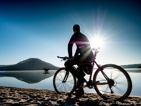 Man biker sit on bike frame and watch the calm lake within sunrise.