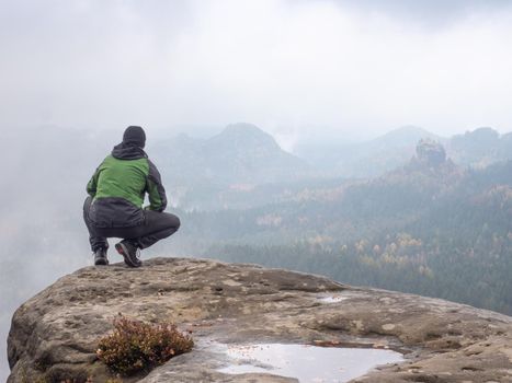 Man sitting at the peak of a mountain on a foggy morning.  Sit squatting on the peak edge and enjoying mountains view valley during heavy mist
