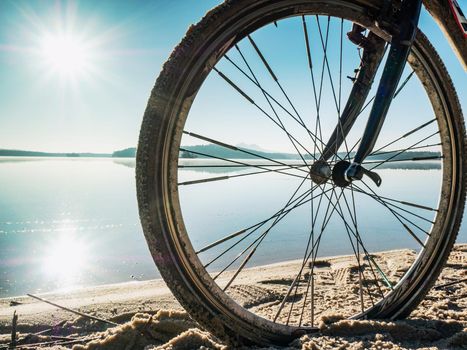 Front wheel of graviel bike in sand of beach. The sun is mirroring in blue water of lake