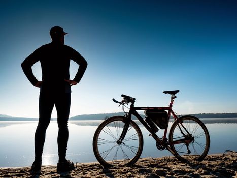 Silhouette of a biker with bike on beach. Rear View of Man Standing with a gravel bike at Sunset. Healthy Lifestyle Concept